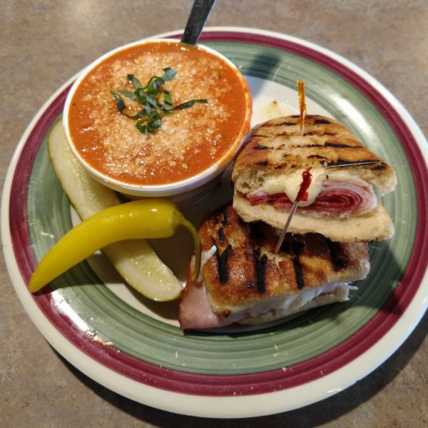 A grilled Italian panini with a side of tomato soup, pickles, and a pepperoncini on a striped plate.
