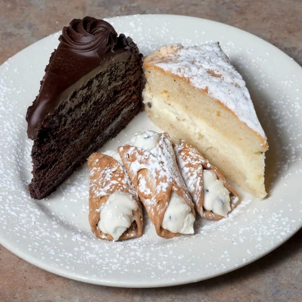A white plate holds a slice of chocolate cake, a slice of cheesecake, and three cannolis, all dusted with powdered sugar.