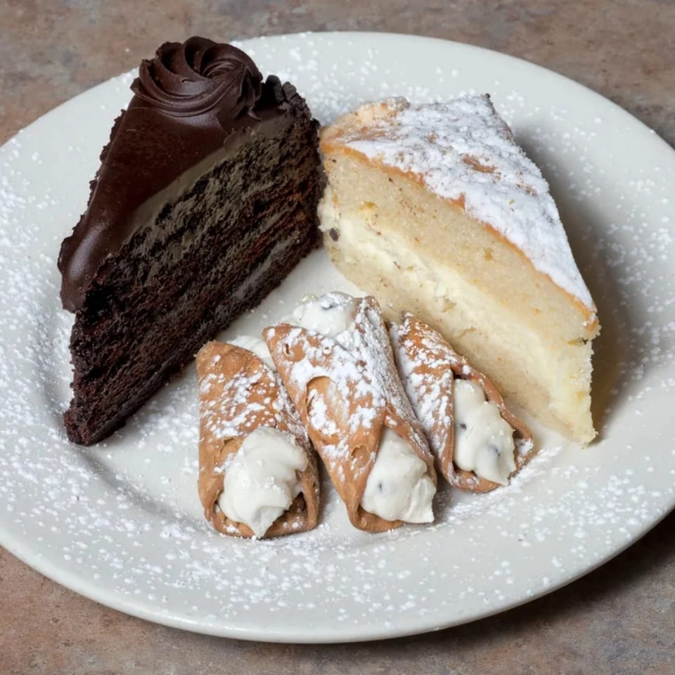 A white plate holds a slice of chocolate cake, a slice of cheesecake, and three cannolis, all dusted with powdered sugar.