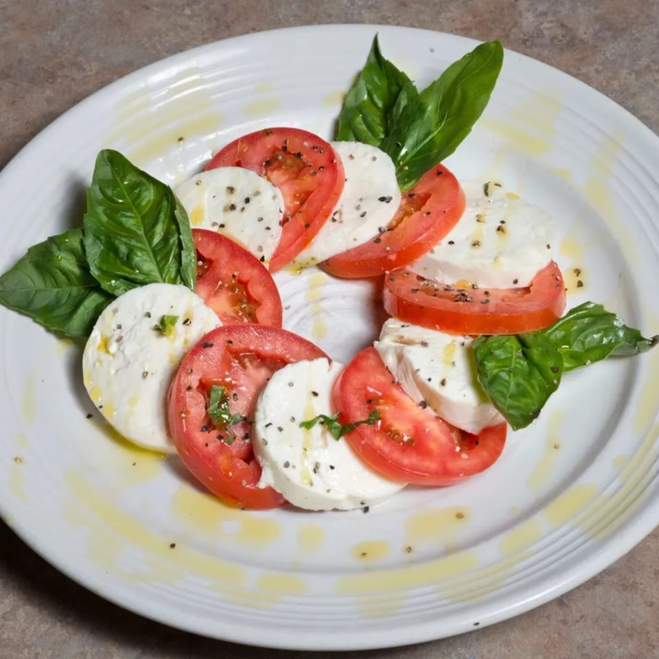 Caprese salad with sliced mozzarella, tomatoes, fresh basil, olive oil, and pepper on a white plate.