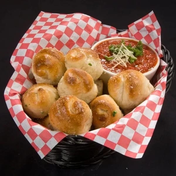 Angelo's West garlic knots in a basket with red and white checkered paper, served with marinara sauce.